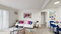 This single-floor unit is designed with a clean, bright aesthetic throughout, characterized by white walls, light wood-style flooring in the main living areas, and an abundance of natural light. The living room serves as the central gathering space, featuring a wall-mounted air conditioning unit, large sliding glass doors dressed with vertical blinds that allow natural light to fill the room, and direct access to an outdoor area. The space is further enhanced by colorful abstract wall art, adding a bold, creative focal point to the otherwise neutral palette.

The galley-style kitchen is efficiently laid out with white cabinetry, tile flooring, a stainless steel sink with a matte black faucet, and white appliances including a coil-top range and refrigerator. Adjacent to the living area, a dedicated media and storage wall provides functional display space.

The bedroom is carpeted for comfort and features a window with outdoor views, a walk-in closet offering generous storage, and its own vibrant wall art that ties the unit's colorful design theme together.

The full bathroom includes a tub-shower combination with white tile surround, a single-sink vanity with white cabinetry, and wood-style flooring that complements the rest of the unit.