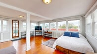 Located at 1133-1137 27th St in San Diego, CA, this thoughtfully designed studio offers a compact yet functional single-floor layout. The open main living area is defined by warm, rich hardwood flooring that stretches throughout, complemented by an abundance of natural light pouring in through large wraparound windows on multiple walls. These windows frame pleasant neighborhood views and give the space an airy, elevated feel. A structural column adds a classic architectural touch to the open-plan living and sleeping area, which flows seamlessly from one zone to the next. The kitchen is a standout feature, outfitted with sage-green shaker-style cabinetry, granite countertops, stainless steel appliances including a gas range and built-in microwave, and a single-basin sink — all finished with light tile flooring. A staircase with a white railing connects the kitchen level to the main living area, adding visual interest and dimension to the layout. The bathroom includes a full tub and shower combination finished with marble-style tile surround. The hallway provides a practical transition between the kitchen and main living space. Access to an outdoor deck area is available through a wood-framed glass door in the living room, extending the livable space outdoors.