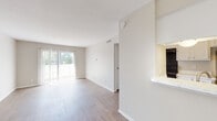 This single-floor unit offers a bright, functional layout with a clean and neutral aesthetic throughout. Light wood-style flooring runs continuously across the main living spaces, creating a cohesive and easy-to-maintain interior. The living area is generously proportioned, benefiting from natural light that streams in through a large sliding glass door fitted with vertical blinds, offering outdoor views and a seamless connection to the exterior. A front entry door opens directly into this open living space, which is anchored by a pendant light fixture overhead and complemented by a built-in pass-through counter that connects to the kitchen.

The galley-style kitchen is efficiently arranged with white cabinetry lining both sides, providing ample storage. Black appliances — including a range, refrigerator, and dishwasher — offer a contrasting accent against the white countertops and cabinetry. A hallway leads from the main living area to the bedroom and bathroom. The bedroom is a quiet, well-proportioned room with a window fitted with horizontal blinds, allowing for natural light and privacy. The bathroom features a full-size tub and shower combination with clean white tile surround, maintaining the unit's consistent neutral palette throughout.