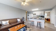 This single-floor unit offers a well-organized layout accommodating three bedrooms and three bathrooms within a compact footprint. The open-concept living and kitchen area forms the central hub of the unit. The living room features wood-look laminate flooring, a ceiling fan with light fixture, large windows with horizontal blinds that allow natural light and outdoor views, and wall-mounted television space. Seating flows naturally into the kitchen area, which is separated by a granite-topped breakfast bar with barstool seating. The kitchen is equipped with white cabinetry, granite countertops, a full suite of white appliances including a refrigerator, electric range, and built-in microwave, as well as a stacked washer and dryer unit conveniently integrated into the space. The three bedrooms feature neutral carpet flooring, ceiling fans, and individual closets with shelving. Each bedroom benefits from direct access to one of the three bathrooms, which include either a walk-in glass-enclosed shower or a shower-tub combination, white vanity cabinetry, and tile flooring. Grey-toned walls and white trim carry consistently throughout, lending the unit a cohesive, clean aesthetic.