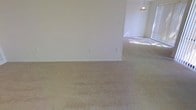 This single-floor space features an open layout with interconnected living areas that flow naturally into one another. Soft, neutral-toned carpet covers the floors throughout, contributing to a warm and understated aesthetic. The walls are finished in a clean, light cream color, creating a bright and airy atmosphere that complements the open feel of the space.

A partial wall divides the main living area from an adjoining room, which benefits from natural light streaming in through a window, brightening the interior. A pendant light fixture hangs in this secondary space, providing a functional focal point overhead. Multiple electrical outlets are positioned along the walls, offering practical convenience throughout the space.

The absence of furniture allows the full scale of the rooms to be appreciated, highlighting the generous proportions of the living areas. The consistent flooring and color palette throughout create a cohesive, unified look. Overall, the space presents a clean, move-in-ready environment with a simple and functional design well-suited to a variety of interior arrangements and personal styles.