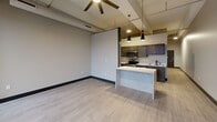 Located at 229 W 3rd St in downtown Davenport, Iowa, this one-bedroom, one-bathroom unit offers a thoughtfully designed layout across a single floor. The open-concept living and kitchen area features light wood-look flooring, a coffered ceiling with ceiling fan, and exposed structural elements that lend an urban, industrial character. The kitchen is appointed with dark cabinetry, stainless steel appliances, and a marble-style peninsula countertop, illuminated by sleek pendant lighting that anchors the space. The spacious bedroom is finished with neutral carpet, bold dark door and baseboard trim, and a ceiling fan, creating a comfortable and cohesive aesthetic. A built-in closet provides convenient storage. The bathroom includes a dark-toned vanity with gold hardware accents and a clean white countertop. A standout feature is the in-unit stacked washer and dryer, conveniently positioned in the hallway near the bathroom. Neutral taupe and gray tones are carried throughout, unified by dark trim work that adds a modern edge. The overall design balances contemporary style with functional, efficient use of space.