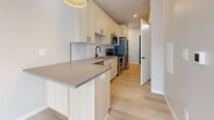 This single-floor residence showcases a thoughtfully designed contemporary interior with a cohesive, minimalist aesthetic. Light-toned hardwood flooring runs throughout the living areas and bedroom, contributing to a bright, airy atmosphere that complements the neutral palette of soft white and greige walls.

The kitchen is a standout feature, equipped with light wood cabinetry extending to the ceiling for generous storage, gray quartz countertops, a white subway tile backsplash, and stainless steel appliances including a range, microwave, and refrigerator. A peninsula with an integrated sink and a matte black faucet provides additional workspace and casual seating potential.

The bedroom opens directly onto a private balcony through expansive floor-to-ceiling sliding glass doors, framing views of the surrounding urban streetscape and filling the room with natural light. The balcony features metal railing and offers a comfortable outdoor extension of the living space.

The bathroom is finished with large-format tile flooring, a light wood vanity with gray countertop, matte black hardware accents, and a full tub-shower combination with crisp white tile surround. The overall design of the space emphasizes clean lines, modern materials, and functional simplicity throughout.