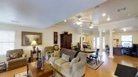 This single-story space presents an open-concept layout that seamlessly connects the living and dining areas, creating a sense of spaciousness and flow. The living area is anchored by a generously sized upholstered sofa and coordinating armchairs, complemented by a warm wood coffee table and layered area rugs that add texture and comfort underfoot. A large dark wood armoire serves as a striking focal point along one wall, accompanied by framed artwork and decorative table lamps that contribute to the space's traditional aesthetic.

Elegant decorative columns with classical detailing define the transition between the living and dining zones without interrupting the open feel of the space. The dining area accommodates a round table with cushioned chairs, ideal for gathering. Recessed lighting and dual ceiling fans with integrated light fixtures ensure consistent illumination and airflow throughout.

Natural light is a notable feature, with French doors at the far end of the space drawing the eye and brightening the interior. The light hardwood flooring runs throughout, tying the areas together cohesively. Overall, the space exudes a warm, classic ambiance with thoughtful architectural details.