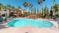 This outdoor amenity area centers around a generously sized freeform swimming pool with crystal-clear turquoise water, creating an inviting resort-like atmosphere. A striking natural rock water feature serves as a focal point within the pool, adding a dramatic, organic element to the design. The pool's curvilinear shape enhances its leisurely, resort-inspired aesthetic.

Adjacent to the main pool is a dedicated spa, offering a more intimate and relaxing water experience. The surrounding pool deck is finished in a warm-toned concrete surface, providing ample space for lounging. Stylish poolside furniture, including chaise lounges and cushioned seating with shaded umbrellas, is thoughtfully arranged to maximize comfort and enjoyment.

The landscaping is a standout feature, with tall fan palms and slender Italian cypress trees creating a lush, layered canopy that frames the space beautifully. Potted plants and additional desert-adapted greenery further enhance the tropical ambiance. The surrounding architecture features warm earth-toned stucco buildings with red tile roofing, complementing the natural desert setting. Abundant natural light and clear skies complete the vibrant outdoor experience.