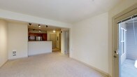 This single-floor residence offers a clean, functional layout designed for comfortable everyday living. The open-plan living area is finished with plush, light-toned wall-to-wall carpeting, creating a warm and inviting atmosphere. Neutral cream-colored walls provide a versatile backdrop throughout the space, allowing for a variety of interior design choices.

The kitchen is seamlessly integrated into the living area and features rich, warm-toned wood cabinetry paired with stainless steel appliances, including a full-sized refrigerator. A raised breakfast bar defines the boundary between the kitchen and the living space, offering a casual dining or workspace option. Track lighting mounted on the kitchen ceiling provides focused, adjustable illumination over the cooking and prep areas, while a recessed ceiling light ensures the entryway zone is well-lit.

The entryway is accessible through a solid door and is flanked by what appears to be a utility or storage panel, adding to the practicality of the space. A secondary interior door nearby suggests access to a closet or additional room. The overall design is straightforward and efficient, prioritizing functionality while maintaining a tidy, well-maintained aesthetic throughout.