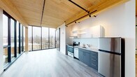 Located at 7373 N Philadelphia Ave in Portland, OR, this thoughtfully designed single-floor unit combines modern aesthetics with warm architectural elements. The space is defined by its striking exposed wood plank ceilings, which add natural warmth and texture, while industrial-style black track lighting and visible structural beams contribute to a contemporary loft-inspired character.

The open main living area is anchored by expansive floor-to-ceiling windows framed in sleek black metal, flooding the interior with abundant natural light and offering elevated views of the surrounding tree-lined neighborhood. A sliding barn door provides a seamless division between spaces while maintaining the open, airy feel.

The kitchen is well-appointed with two-tone cabinetry — charcoal lower cabinets paired with white upper shelving — and a full suite of stainless steel appliances including a range, dishwasher, and refrigerator. Light gray wood-tone flooring runs throughout the main living area, complementing the overall clean, modern palette.

The bathroom features a dark-toned vanity with a white countertop, an integrated sink, a large mirror, and a full-size soaking tub with shower combination. A light wood-tone hallway with clean white walls leads from the entry door, providing a welcoming transition into the living space.