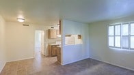This single-story space offers a practical and functional layout designed for comfortable everyday living. The open floor plan creates a natural connection between the living area and the kitchen, with a defining architectural feature — a partial dividing wall with a pass-through window — providing visual separation while maintaining an open feel throughout.

The kitchen is equipped with warm-toned wood cabinetry that adds a natural, inviting element to the space. White appliances, including a range and refrigerator, are integrated neatly into the layout, complementing the light-colored countertops. The kitchen floor is finished with tile, offering durability and ease of maintenance in a high-use area.

Transitioning into the living area, the flooring shifts to carpet, adding warmth and comfort underfoot. The walls throughout are painted in neutral tones, creating a clean and versatile backdrop suitable for a variety of décor styles. A doorway visible in the background leads to additional rooms, suggesting a well-connected and accessible floor plan. The overall ambiance is light, airy, and move-in ready.