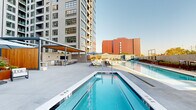 This thoughtfully designed outdoor amenity space offers a comprehensive array of leisure zones set against the backdrop of a modern high-rise building. At the heart of the space is a generously sized lap pool with crystal-clear water, lined with rows of lounge chairs and shaded umbrellas for relaxed sunbathing. Adjacent to the pool, a dedicated spa with stainless steel entry rails provides an intimate retreat. A separate courtyard zone features a lush, well-manicured lawn framed by raised Corten steel planters brimming with ornamental grasses, shrubs, and seasonal plantings, creating a verdant, park-like atmosphere. An inviting fire pit seating area with plush, navy-blue lounge chairs and string lights overhead adds warmth and ambiance for evening gatherings. Outdoor dining tables with umbrellas are arranged nearby for al fresco meals. A rooftop terrace level offers expansive sectional seating with sweeping views of the surrounding urban skyline, complemented by additional raised planters. Throughout, the design integrates clean-lined modern architecture with natural materials, open sky exposure, and layered landscaping to create a vibrant and functional outdoor living environment.
