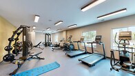 Located at 353 Vine St in West Lafayette, Indiana, this fitness center offers a comprehensive, single-floor workout facility designed to support a wide range of exercise routines. The space is thoughtfully laid out to maximize functionality, with a clean, open floor plan that allows users to move freely between equipment zones.

The cardio area is prominently featured and includes multiple treadmills, an elliptical trainer, and a rowing machine, all positioned near windows that bring in natural light and provide views of the surrounding greenery. Overhead, a series of flush-mounted fluorescent lighting fixtures ensures the space is consistently well-lit throughout.

Strength training is well-represented with a multi-station cable machine and a dedicated free weights area housing a full rack of hex dumbbells in varying sizes. An adjustable weight bench adds further versatility to the strength zone. On the opposite end of the room, a vibrant functional training corner features resistance bands, medicine balls, stability balls in assorted sizes, and yoga/exercise mats for stretching and floor-based workouts.

The interior finishes are clean and modern, with neutral-toned walls accented by warm yellow paint in the functional training area, durable speckled flooring, and white ceilings. The space is accessible via multiple entry points, including a dedicated exit door, making it practical and easy to navigate for daily use.