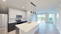 This well-appointed single-floor unit offers a thoughtfully designed open-concept layout encompassing a living room, kitchen, bedroom, closet, and full bathroom. The kitchen serves as a central highlight, featuring a large waterfall-edge island with an undermount sink, white quartz countertops, white upper cabinetry contrasted by dark lower cabinets, a classic subway tile backsplash, and a full suite of stainless steel appliances. Three modern pendant lights with gold accents hang above the island, adding a stylish focal point. Light wood-look flooring flows throughout the main living areas, enhancing the bright, airy atmosphere. The living room opens directly to a private balcony through sliding glass doors, offering outdoor views of surrounding greenery and the neighborhood. The bedroom features plush carpeting, a large window with natural views, and direct access to a dedicated closet. The full bathroom is finished with a dark wood vanity, white quartz countertop, large mirror, and a glass-enclosed walk-in shower, complemented by light marble-style tile flooring. Recessed lighting and neutral wall tones contribute to the clean, contemporary ambiance throughout.