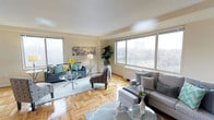 This single-floor space presents a well-appointed living area characterized by classic parquet hardwood flooring that adds warmth and texture throughout. Large windows on multiple walls flood the room with abundant natural light while offering views of the surrounding landscape, including mature trees and neighbouring structures. The textured ceiling and neutral cream-toned walls provide a clean, understated backdrop that complements a variety of décor styles. The living area is generously proportioned, comfortably accommodating multiple seating arrangements, including sofas, accent chairs, and glass-topped coffee tables. The layout encourages an open, airy feel, enhanced by the corner window configuration that maximizes daylight exposure from more than one direction. Decorative touches such as wall art, indoor plants, and layered area rugs contribute to the overall livable ambiance. The space benefits from baseboard heating positioned beneath the windows, a practical feature consistent with the building's construction. Overall, the living area strikes a balance between functional design and comfortable aesthetics, making it a versatile and welcoming environment.