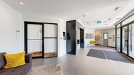 Located at 235 Stillwater Dr in Saskatoon, Saskatchewan, this building lobby presents a clean, contemporary aesthetic designed to make a strong first impression. The single-floor space is anchored by a striking dark-clad fireplace column that serves as a bold architectural focal point, dividing the entry vestibule from the main lounge area. Large-format light grey tiles extend across the entire floor, lending the space a cohesive and polished look. A set of floor-to-ceiling black-framed glass doors forms the building entrance, flooding the interior with natural light and offering views of the surrounding greenery. The lounge area is furnished with a charcoal sofa accented by yellow throw pillows, complemented by a pair of vibrant yellow accent chairs that add a pop of colour against the white walls. Framed artwork lines one wall, contributing to the refined atmosphere. Practical amenities include a parcel locker station and a residential mailbox unit, ensuring convenience for building occupants. Recessed ceiling lighting and a tray ceiling detail above the entrance zone add architectural depth, while grey entry mats provide a practical transition from the outdoors. The overall ambiance is modern, bright, and welcoming.