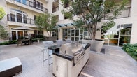 Located at 5670 W Wilshire Blvd in the heart of Los Angeles, this beautifully designed outdoor courtyard offers a well-appointed communal amenity space ideal for gathering and relaxation. The centerpiece of the courtyard is a full-size stainless steel gas BBQ grill station, complete with a black granite countertop, built-in storage, and bar-height seating — perfect for outdoor entertaining. Surrounding the grill area, the courtyard opens into a spacious patio furnished with wicker lounge seating, coffee tables, and an umbrella-shaded dining set, creating a comfortable and inviting atmosphere. Decorative raised planters filled with polished black river rock and mature trees add a sophisticated, resort-like aesthetic, while lush greenery, manicured hedgerows, and verdant shrubs line the perimeter, providing a natural sense of privacy and tranquility. String lights overhead enhance the ambiance for evening use. The courtyard is enclosed by the surrounding multi-story building, with upper-level balconies overlooking the space. Glass-paneled entry doors and dark wood slatted privacy screens contribute to the refined architectural character of the space. A glimpse of a pool or spa area is visible nearby, rounding out this thoughtfully designed outdoor retreat in the Wilshire corridor.