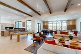 Located in Peoria, Arizona, this single-floor community clubhouse offers a generously sized, open-concept living area designed for both relaxation and socializing. The space is anchored by striking exposed wood ceiling beams that add warmth and architectural character to the otherwise contemporary interior. Large-format light tile flooring flows throughout the main lounge area, while a contrasting dark tile section defines a separate seating zone, creating distinct areas within the open layout.

A dramatic floor-to-ceiling wood-paneled feature wall serves as the focal point of the lounge, housing a linear marble-surround fireplace flanked by dual flat-screen televisions and built-in shelving. Plush sofas and accent chairs in neutral tones are accented with bold terracotta and rust-colored throw pillows, lending a warm, Southwest-inspired palette to the space.

A full-size pool table takes center stage in the recreational area, positioned beneath recessed lighting for an inviting game-room atmosphere. A wet bar with counter seating is visible along one wall, ideal for entertaining. A row of elegant arched windows along one side of the room floods the interior with natural light while offering views of the outdoor surroundings. Black-framed glass doors provide seamless access to adjacent areas and the exterior, enhancing the sense of openness throughout.