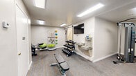 Located at 3555 N First Ave in Tucson, Arizona, this fitness center offers a thoughtfully organized exercise space across a single floor. The room features neutral white walls, patterned carpet flooring, and a combination of recessed and flush-panel overhead lighting that keeps the space bright and functional. A ceiling fan in the cardio area provides additional air circulation for comfort during workouts.

The cardio section is equipped with multiple Spirit treadmills, an elliptical trainer, and a stationary recumbent bike, positioned near a window that allows natural light to filter in. Strength training is well-represented with a multi-function cable machine, a leg extension/curl machine, a dedicated free weight rack holding a full set of dumbbells, and an adjustable workout bench. A wall-mounted accessory rack holds resistance bands, medicine balls, and stability balls, catering to a variety of fitness routines.

A wall-mounted flat-screen television adds entertainment value, while a dual-basin water fountain station ensures easy hydration access. A charming barn-style sliding door adds a subtle rustic design element to the otherwise modern interior. The space is accessed via a glass exterior entry door and includes a secondary interior door, making it conveniently accessible. This fitness center provides a comprehensive and comfortable environment for a full range of exercise activities.
