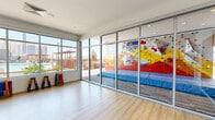 Located at 1100 East Apache Boulevard in Tempe, Arizona, this contemporary fitness studio offers a well-appointed, open-plan layout designed to support a variety of exercise and wellness activities. The space features light hardwood flooring throughout, contributing to a clean and energizing atmosphere. A dramatic, large-scale photographic mural of a sweeping canyon landscape dominates one wall, adding a bold visual focal point and an element of natural inspiration to the interior. A mounted display screen is integrated into this feature wall, offering functional utility for instruction or entertainment. Floor-to-ceiling glass walls and expansive windows flood the studio with abundant natural light and provide unobstructed views of the outdoor amenity area, which includes a vibrant bouldering wall with colorful holds and crash pads, as well as a pool. The glass partition wall creates a seamless visual connection between the interior studio and the outdoor climbing area. A full-length mirror along one wall further enhances the sense of openness and supports fitness use. Fitness accessories, including dumbbells, resistance bands, and exercise mats, are neatly stored along the perimeter. Recessed lighting and modern HVAC fixtures in the white ceiling complete the polished, functional design of this active amenity space.