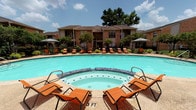 This community's outdoor amenity area is thoughtfully designed to offer a comprehensive resort-style experience. At its centerpiece is a generously sized swimming pool with clear turquoise water, complemented by an adjacent circular spa finished with decorative tile detailing. The pool deck is furnished with orange lounge chairs and market umbrellas, providing ample space for sunbathing and relaxation.

A covered patio extends alongside the pool area, offering a shaded retreat equipped with a ceiling fan and comfortable striped lounge seating arranged around decorative side tables. Large potted plants and greenery are strategically placed throughout, adding natural color and dimension to the space. An outdoor bar-height counter with seating and a built-in grill station provide a dedicated space for outdoor entertaining and dining.

Access to the pool area is provided through bold red French doors framed by brick exteriors, accented with potted flowering plants that enhance the entryway's curb appeal. The surrounding brick residential buildings with pitched rooflines create an enclosed, courtyard-like atmosphere. The entire outdoor space is secured with wrought iron fencing, providing both safety and an open, airy feel. Mature trees visible above the rooflines contribute additional shade and a lush, established character to the overall environment.