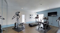 This single-story fitness room in Irving offers a clean, functional space designed for residents to maintain an active lifestyle. The room features a light, neutral color scheme with white upper walls and a calming blue accent along the lower portion, creating a fresh and motivating atmosphere. Wood-look flooring runs throughout, providing a durable and easy-to-maintain surface beneath the equipment.

The space is equipped with a multi-station cable weight machine for strength training, multiple treadmills, and an elliptical trainer, giving users a well-rounded selection of cardio and resistance options. A wall-mounted monitor is available for entertainment during workouts, and a fan is positioned to provide additional airflow.

Natural light enters through windows fitted with horizontal blinds, keeping the space bright while allowing for adjustable privacy. Recessed lighting in the ceiling supplements the natural light for early morning or evening workouts. A glass-paneled door provides direct exterior access, enhancing ventilation and convenience. Overall, the room is thoughtfully arranged to maximize usability within a comfortable, inviting environment.