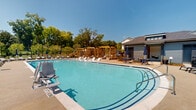 Located at 1300 S Davis Rd in LaGrange, Georgia, this outdoor aquatic amenity area is designed to deliver a resort-style recreational experience. The centerpiece is a generously sized swimming pool with crystal-clear blue water, gently curved edges, and accessible entry points including a pool lift, ensuring usability for all residents. The pool is surrounded by a broad concrete deck that accommodates an extensive collection of chaise lounge chairs and umbrella-shaded seating, providing ample space for sunbathing and relaxation.

A standout architectural feature of the space is a series of warm-toned wooden pergola structures positioned along one side of the pool, offering shaded gathering areas with a stylish, contemporary aesthetic. Adjacent to the pool, a single-story pool house finished in cool gray siding provides practical amenities and serves as a functional anchor to the overall layout. Outdoor dining tables and chairs are arranged near the pool house, creating an inviting social setting.

The surrounding landscape is lush with mature trees that provide a natural backdrop and additional shade, while open green space and clear sightlines beyond the perimeter fencing contribute to an airy, open ambiance. The entire area is enclosed by black metal fencing, ensuring privacy and security throughout this well-appointed outdoor amenity space.