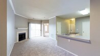 This single-story space showcases an open-concept layout that seamlessly connects the living area and kitchen. The carpeted living room is a generous size, anchored by a tile-surround fireplace with a white mantle along one wall. Crown molding runs throughout the main living area, adding a refined architectural detail to the otherwise neutral, gray-toned walls and white ceilings.

Natural light enters through a large sliding glass door that leads to a balcony, offering an outdoor extension of the living space. Adjacent to the living room, a doorway leads to an additional room, visible in the background, which features its own window and closet doors, suggesting a bedroom or secondary living space.

The kitchen is defined by a wraparound peninsula with a light granite-style countertop and an integrated sink. Dark countertops are also visible along the perimeter cabinetry. A large mirror in the kitchen area reflects the entryway, which features two sets of double doors — likely a coat closet and the main entrance — enhancing the sense of depth and light throughout the space.