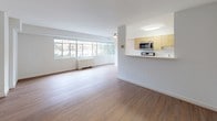 This single-floor residence is thoughtfully designed with a clean, modern aesthetic and a functional layout. The galley-style kitchen is a standout feature, outfitted with light wood cabinetry extending from floor to ceiling, white quartz countertops, stainless steel appliances including a gas range, microwave, refrigerator, and dishwasher, and white tile flooring. A pass-through counter connects the kitchen to the main living area, enhancing the open feel of the space.

The living area is generously proportioned, featuring warm wood-tone laminate flooring and large windows that admit abundant natural light and provide views of surrounding greenery. A radiator unit runs beneath the windows, and the white walls contribute to a bright, airy atmosphere throughout.

A bedroom features wood-tone flooring consistent with the rest of the unit and includes a sliding-door closet that houses an in-unit washer/dryer combination unit, adding considerable convenience.

The bathroom is finished with white subway-style wall tiles and includes a full soaking tub with an overhead shower and chrome fixtures. The overall design of the space prioritizes clean lines, neutral tones, and practical functionality.