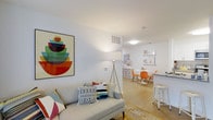 This single-floor residence at Eagle Rock at Freehold offers an open, airy layout designed for comfortable everyday living. The living area is anchored by a light gray sofa accented with vibrant throw pillows, complemented by a sleek tripod floor lamp that adds a contemporary touch. A bold, colorful piece of wall art brings energy and personality to the space, setting a playful yet modern tone throughout. The clean white walls and light-toned flooring work together to maximize the sense of brightness and openness. The living space flows naturally into the dining area, where a round white table is paired with modern molded chairs in a warm orange hue, creating a cheerful and functional dining setting. Floating shelves display curated artwork and décor, adding visual interest without overwhelming the space. A countertop bar area with a white stool provides an additional casual seating option, ideal for quick meals or entertaining. Ceiling-mounted lighting and HVAC vents are neatly integrated into the white ceiling, maintaining the clean, uncluttered aesthetic. Overall, this residence combines a practical, open floor plan with a bright, contemporary design sensibility that feels both welcoming and stylish.