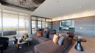 This elegantly designed high-rise space offers a seamless blend of contemporary luxury and thoughtful functionality. The main living area commands attention with its dramatic artistic ceiling mural, a large linear fireplace set against a textured wood-clad accent wall, and a mounted flat-screen television. Generous sectional seating in rich earth tones anchors the space, while a steel-framed glass partition wall adds an architectural layer of visual depth, dividing zones without sacrificing the sense of openness.

Floor-to-ceiling glass walls flood the interior with abundant natural light and frame sweeping panoramic city views that extend to the horizon. A spacious wraparound terrace offers an exceptional outdoor living area, furnished with plush white seating and lounge chairs, perfectly positioned to take in the elevated cityscape.

The dining area features a solid wood table paired with sculpted upholstered chairs beneath a striking multi-arm brass and black chandelier. A dark accent wall with built-in shelving and curated artwork creates an intimate backdrop for the dining experience.

An additional lounge area showcases a dramatic marble-top coffee table, deep velvet sofas, and swivel armchairs, all oriented toward the expansive glazed facade. Elsewhere, a bar-height counter with richly grained wood and a dark stone dining table with white shell chairs provide versatile entertaining options. The overall ambiance is refined, cosmopolitan, and sophisticated.