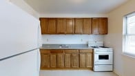 This single-floor space includes a well-appointed kitchen that serves as a central feature of the layout. The kitchen is designed with warm-toned, honey-brown wood cabinetry that extends both above and below the countertops, providing generous storage capacity. Upper cabinets reach toward the ceiling, maximizing vertical storage space, while the lower cabinets offer equally ample room for organization.

The countertops feature a speckled granite-style surface that complements the wood tones of the cabinetry, running the full length of the kitchen wall and incorporating a stainless steel double-basin sink. A white subway tile backsplash lines the wall between the upper and lower cabinets, adding a clean, classic aesthetic to the space.

White appliances complete the kitchen, including a gas range with multiple burners and a full-sized refrigerator positioned at the end of the counter run. The light-toned flooring and neutral walls contribute to a bright, clean atmosphere throughout. The overall design of the kitchen emphasizes practicality and functionality, with a cohesive, traditional aesthetic defined by its warm wood tones and classic finishes.