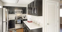 Located at 81 Charleston Sq in Johnson City, this two-floor home offers a practical and well-organized layout with distinct living zones spread across its two levels. The floor plan reveals an open arrangement on the main level, where the living and kitchen areas connect to create a cohesive common space.

The kitchen is a standout feature, equipped with dark espresso cabinetry that contrasts sharply against light granite-style countertops. Stainless steel appliances, including a range and over-the-range microwave, contribute to the kitchen's functional and modern character. Ample upper cabinetry provides generous storage throughout.

Hardwood-style flooring runs throughout much of the main level, lending warmth and visual continuity to the space. A hallway connects the common areas to additional rooms, lined with neutral gray walls and white trim that create a clean, understated aesthetic. Textured ceilings and visible HVAC venting are consistent throughout.

Natural light filters in through windows at the end of the hallway, brightening the interior and reinforcing the home's airy feel. The staircase leads to the second floor, extending the overall living space and offering additional room configurations above the main level.