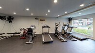 Located at 10010 Kempwood Dr in Houston, Texas, this dedicated fitness center offers a well-equipped, single-floor space designed to support a full range of workout routines. The room features durable rubber flooring throughout, providing a safe and functional surface for exercise. Recessed lighting illuminates the space evenly from above, complemented by natural light streaming in through large windows that offer views of the surrounding outdoor area.

The cardio section is outfitted with multiple machines, including treadmills, elliptical trainers, and a stationary cycle, all arranged to allow comfortable use. On the strength training side, a cable machine with weight stacks serves as a centerpiece, accompanied by a weight bench and free weights. Floor-to-ceiling mirrors line one wall, visually expanding the space and allowing users to monitor their form during workouts. Wall-mounted fans provide ventilation throughout the room.

Additional amenities include built-in wall shelving for personal items, a posted fitness center rules sign, and a fire extinguisher for safety. A glass-paneled door provides direct access to the exterior courtyard, further connecting the space to the broader property. The overall atmosphere is clean, functional, and well-maintained, making it a practical resource for fitness-focused residents.