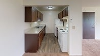 This single-floor unit at Rosewood in Inkster, Michigan, offers a straightforward and functional layout across two bedrooms and one bathroom. The living area is spacious and carpeted, featuring a chandelier overhead and a large window that draws in natural light and provides views of the surrounding neighborhood. The front entry door opens directly into this open main living space, which connects seamlessly to the kitchen area.

The kitchen is designed as a galley-style space, equipped with dark wood cabinetry on both sides providing ample storage, light-toned countertops, an integrated sink, a white electric range, and a refrigerator. Wood-look flooring runs throughout the kitchen, contrasting with the carpeting in the rest of the unit.

Both bedrooms are carpeted and finished with neutral white walls. Each room benefits from windows that allow natural light to filter in, with one bedroom featuring dual windows offering views of the surrounding greenery. Overhead lighting and ceiling vents are present in the bedrooms.

The bathroom includes a full tub and shower combination with white tile surround, a vanity, and distinctive multicolored mosaic tile flooring, adding a classic touch to the space. Overall, the unit presents a clean, neutral palette suitable for comfortable everyday living.
