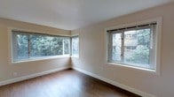 This single-floor space presents a clean, minimalist aesthetic defined by warm hardwood flooring and a neutral, light beige color palette across its walls and ceiling. The room showcased is generously lit by natural light, courtesy of multiple windows positioned along two walls. A wide, multi-pane corner window anchors one side of the room, framing views of surrounding greenery and trees, while a second casement-style window on the adjacent wall offers additional outdoor sightlines toward neighboring architecture and foliage. Together, these windows ensure a bright, well-ventilated environment throughout the day. The smooth, white ceiling and crisp white baseboards complement the warm tones of the hardwood floor, contributing to a cohesive and polished interior. The open layout of the room provides a versatile and uncluttered living area, well-suited to a variety of furniture arrangements. The overall ambiance is calm and inviting, with an emphasis on natural light and a connection to the outdoors, making it a functional and comfortable space.