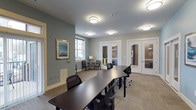 This single-floor professional space is thoughtfully designed to support a range of collaborative and private work needs. The main area centers around a large, curved dark-wood conference table surrounded by ergonomic mesh chairs, making it well-suited for group meetings or focused work sessions. Neutral-toned carpet with a subtle linear pattern runs throughout, complementing the soft blue-gray and warm white wall palette that lends the space a calm, professional atmosphere. Flush-mounted ceiling fixtures provide consistent, even lighting across the room. Natural light filters in through a set of windows, adding brightness to the conference area. Framed artwork adorns the walls, contributing a polished finishing touch. At the rear of the space, a pair of glass-paneled French doors leads to a private enclosed room, offering a more secluded setting for confidential meetings or focused work. A casual seating nook with armchairs and a floor lamp is tucked near the windows, providing a more relaxed option for informal conversations. The overall design balances functionality with a welcoming, well-appointed aesthetic suited for a professional or community-oriented environment.