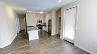 This single-floor residence offers a thoughtfully designed open-concept layout that seamlessly connects the kitchen and living areas, creating a bright and functional living environment. The contemporary kitchen is a standout feature, equipped with stainless steel appliances including a refrigerator, gas range, and built-in microwave. Crisp white cabinetry with dark countertops provides ample storage and workspace, while a central kitchen island with an integrated sink adds both convenience and a natural gathering point. The flooring throughout features warm hardwood-style planks that complement the neutral, light-toned walls, contributing to a clean and cohesive aesthetic. Recessed and flush-mount ceiling lighting ensures the space is well-lit throughout. A glass-paneled door with slatted blinds leads to an outdoor area, allowing for natural light to filter into the space. A hallway visible from the main area suggests additional rooms beyond, offering a sense of depth and privacy. Overall, the space presents a modern, well-appointed interior with a practical and inviting atmosphere suited for everyday living.