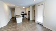 This single-floor residence offers a thoughtfully designed open-concept layout that seamlessly connects the kitchen and living areas, creating a bright and functional living environment. The contemporary kitchen is a standout feature, equipped with stainless steel appliances including a refrigerator, gas range, and built-in microwave. Crisp white cabinetry with dark countertops provides ample storage and workspace, while a central kitchen island with an integrated sink adds both convenience and a natural gathering point. The flooring throughout features warm hardwood-style planks that complement the neutral, light-toned walls, contributing to a clean and cohesive aesthetic. Recessed and flush-mount ceiling lighting ensures the space is well-lit throughout. A glass-paneled door with slatted blinds leads to an outdoor area, allowing for natural light to filter into the space. A hallway visible from the main area suggests additional rooms beyond, offering a sense of depth and privacy. Overall, the space presents a modern, well-appointed interior with a practical and inviting atmosphere suited for everyday living.