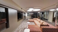 This single-floor space is designed as a contemporary lounge or communal gathering area, offering a polished and welcoming atmosphere. The room is anchored by a large, L-shaped sectional sofa upholstered in a striking geometric, warm-toned pattern, complemented by small round accent tables that provide functional surfaces throughout the seating area. A wall-mounted flat-screen television is recessed into a dedicated niche along one wall, making it a natural focal point for the space.

The flooring features a layered approach, combining patterned carpeting that adds visual depth and texture underfoot. Overhead, a cluster of large, circular flush-mount light fixtures provides bright, even illumination across the main seating area, contributing to the modern aesthetic.

The space extends into additional areas visible in the background, including what appears to be a glassed-in room and a secondary zone with mirrored surfaces and additional seating or equipment, suggesting a multifunctional layout. Slim floor lamps and carefully placed accent lighting further enhance the ambiance.

The overall design balances bold, retro-inspired décor elements with clean architectural lines, creating a space that is both visually engaging and practically arranged for social interaction or relaxed gatherings.
