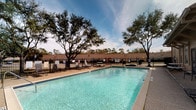 This community amenity space combines an inviting outdoor pool area with a well-designed single-story clubhouse interior. The outdoor pool features clear, blue-tinted water set within a generously sized rectangular pool, surrounded by a paved deck equipped with lounge chairs and seating. Mature trees frame the space, providing natural shade, while a wrought iron perimeter fence ensures safety and security. The open sky above creates a bright, airy atmosphere ideal for outdoor relaxation.

The clubhouse interior is thoughtfully designed with high ceilings adorned with crown molding, recessed lighting, and a striking multi-arm chandelier that serves as a focal point. Dark hardwood-style flooring runs throughout, complementing the crisp white walls. The main gathering area features an open layout with comfortable, modern furnishings, including a tan leather accent chair and patterned area rugs, creating an inviting lounge atmosphere. Large windows and French doors allow abundant natural light to filter throughout the space.

An adjacent room serves as a functional office or business area, featuring tall ceilings, multiple windows, decorative artwork, and a multifunction printer station. The entryway includes a glass exterior door that allows additional natural light, a small credenza, and decorative wall accents, completing the polished, welcoming character of this community facility.