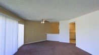 This single-floor space features an open-concept living area finished with dark wall-to-wall carpeting that grounds the room with a warm, understated tone. The walls are painted in a muted tan hue, contributing to a neutral and functional ambiance throughout. A textured popcorn ceiling adds a classic touch overhead, with a ceiling fan centrally positioned to provide air circulation and supplemental lighting across the main living space.

A half-wall pass-through connects the living area to the kitchen, creating a sense of openness between the two spaces while maintaining a defined layout. The kitchen, visible through both the pass-through and a doorway opening, is outfitted with white cabinetry and appears to offer a practical workspace with adequate storage. The cabinetry extends to the upper portions of the kitchen walls, maximizing vertical storage potential.

Natural light enters the living area through a window along one wall, brightening the space during daylight hours. The overall layout is straightforward and efficient, with clean wall lines and a clear flow between the living and kitchen areas, making the space adaptable to a variety of furnishing arrangements.