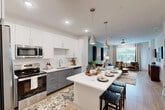 Located at 6735 Standifer Gap Rd in Chattanooga, TN, this single-floor, 2-bedroom, 2-bathroom residence offers a thoughtfully designed open-concept layout that maximizes its comfortable footprint. The kitchen is a standout feature, showcasing two-tone cabinetry with crisp white upper cabinets and slate gray lower cabinets, a classic subway tile backsplash, stainless steel appliances, and a generous island with pendant lighting — perfect for dining and entertaining. Light wood-tone flooring runs throughout, creating a seamless flow into the living room, which is bathed in natural light from large windows with outdoor views and features a ceiling fan and direct access to a private exterior door. The living area is accented by a soft teal feature wall, adding a pop of color to the bright, neutral palette. The primary bedroom offers a cozy retreat with plush carpet, a bold dark accent wall, a ceiling fan, and a large window overlooking the surrounding greenery. The bathrooms are finished with clean white walls, wood-look flooring, glass-enclosed showers with marble-style tile, and gray vanities with white countertops. Two closets provide ample storage throughout the residence, completing this modern and functional Chattanooga home.