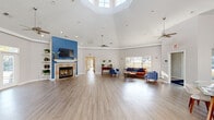 This single-floor community clubhouse is designed to impress with its dramatic vaulted and tray ceilings that lend an expansive, airy atmosphere throughout. The main gathering area serves as the centerpiece of the space, anchored by a tile-surround fireplace set against a bold blue accent wall with a mounted flat-screen television and built-in shelving. Ceiling fans keep the open-plan layout comfortable, while recessed lighting and a striking skylight flood the interior with natural light. Light-toned hardwood-style flooring flows seamlessly through every area, unifying the space visually. A generously sized lounge area offers comfortable seating with a mix of vibrant blue and orange upholstered furniture, creating an inviting and modern aesthetic. The dining area features a glass-top table with contemporary chairs positioned beneath soaring ceilings, with large windows providing views of the surrounding exterior. The fully equipped kitchen is a standout, boasting bold blue walls, white cabinetry, stone-look countertops, black appliances, and a wraparound bar with bar stool seating illuminated by pendant lighting. Multiple exits lead to outdoor areas, and a connecting hallway provides access to additional rooms. The overall ambiance is bright, welcoming, and thoughtfully designed for community gatherings.