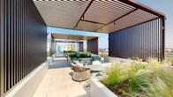 This expansive rooftop terrace offers a well-designed outdoor retreat elevated above the surrounding cityscape. The space is organized into distinct zones, each serving a specific function while maintaining a cohesive contemporary aesthetic. Overhead, slatted wood pergolas provide partial shade across multiple seating areas, casting filtered light onto the light-toned stone pavers below. Dark vertical cladding panels define the structural columns and enclosures, creating visual contrast against the bright, open sky.

Generously sized raised planters filled with ornamental grasses and flowering plants are integrated throughout, adding texture, greenery, and natural separation between zones. Multiple lounge configurations feature modern outdoor sofas and chairs in neutral tones accented with bold yellow and patterned cushions, arranged around low tables to encourage gathering and conversation.

A covered outdoor kitchen station is equipped with a wall-mounted television, built-in cabinetry with warm wood accents, a sink, and counter space, making it well-suited for entertaining. Glass railings along the perimeter allow unobstructed panoramic views of the surrounding urban skyline. The overall ambiance is modern and polished, balancing architecture, landscaping, and amenities into a cohesive, functional rooftop environment.