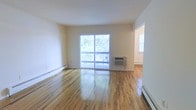 This single-floor residence offers a clean, functional layout highlighted by warm, polished hardwood floors that run throughout the main living area. The living room is a standout space, bathed in natural light streaming through a large sliding glass door that opens onto a private balcony overlooking a lush, green tree canopy — providing a pleasant outdoor connection and scenic views. The bright, neutral white walls amplify the sense of openness and allow for versatile interior design possibilities. A wall-mounted air conditioning unit ensures comfortable climate control during warmer months, while baseboard heating units along the perimeter provide warmth in cooler seasons. An open passage leads to an adjoining area, which appears to benefit from its own window, adding further natural light to the space. The overall ambiance is airy, simple, and move-in ready, with the gleaming hardwood floors serving as the most distinctive design element throughout. The single-floor layout offers convenient, step-free living, making the space practical and accessible.