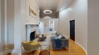 Located at 37 Buena Pl in Rochester, NY, this single-floor space is a striking example of historic architecture thoughtfully updated with contemporary touches. The layout spans a generous footprint, with interconnected rooms flowing through elegant arched doorways supported by decorative columns and intricate plasterwork moldings.

The main living area commands attention with its soaring ceiling height, elaborate crown molding, and a grand marble fireplace featuring ornately carved surrounds. A circular statement light fixture anchors the room, while warm hardwood flooring runs throughout. The adjacent bedroom benefits from dramatic ceiling height, arched windows that draw in abundant natural light, and detailed plaster cornices that echo the classical character found throughout the space.

A kitchenette showcases dark marble-look countertops, a stainless steel undermount sink, white shaker cabinetry, and a bold black-and-white hexagonal tile backsplash. The bathroom offers a clean, modern aesthetic with a frameless glass walk-in shower and neutral tile finishes.

Throughout, the space masterfully balances preserved historic craftsmanship—ornamental pilasters, decorative friezes, and period moldings—with refined modern furnishings and fixtures, resulting in a sophisticated and distinctive interior.