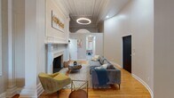 Located at 37 Buena Pl in Rochester, NY, this single-floor space is a striking example of historic architecture thoughtfully updated with contemporary touches. The layout spans a generous footprint, with interconnected rooms flowing through elegant arched doorways supported by decorative columns and intricate plasterwork moldings.

The main living area commands attention with its soaring ceiling height, elaborate crown molding, and a grand marble fireplace featuring ornately carved surrounds. A circular statement light fixture anchors the room, while warm hardwood flooring runs throughout. The adjacent bedroom benefits from dramatic ceiling height, arched windows that draw in abundant natural light, and detailed plaster cornices that echo the classical character found throughout the space.

A kitchenette showcases dark marble-look countertops, a stainless steel undermount sink, white shaker cabinetry, and a bold black-and-white hexagonal tile backsplash. The bathroom offers a clean, modern aesthetic with a frameless glass walk-in shower and neutral tile finishes.

Throughout, the space masterfully balances preserved historic craftsmanship—ornamental pilasters, decorative friezes, and period moldings—with refined modern furnishings and fixtures, resulting in a sophisticated and distinctive interior.
