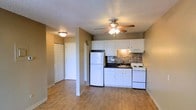 This single-floor space showcases a practical and well-appointed open layout. The kitchen serves as a central feature, equipped with white cabinetry, dark countertops, and a mosaic tile backsplash that adds a contemporary touch. White appliances, including a full-size refrigerator and electric range with overhead ventilation, provide full functionality. A stainless steel sink completes the kitchen setup.

The wood-look laminate flooring runs throughout the space, offering a warm and cohesive aesthetic that ties the rooms together. A ceiling fan with integrated lighting is centrally positioned, ensuring both air circulation and ambient illumination. An additional flush-mount light fixture provides supplemental lighting toward the hallway.

A hallway visible from the main living area leads to additional areas of the unit, including what appears to be a closet with bi-fold doors, offering practical storage solutions. The textured ceiling adds a classic residential feel, while the neutral tan wall tones create a versatile backdrop suitable for a variety of décor styles. Overall, the space presents a clean, functional, and move-in ready environment.