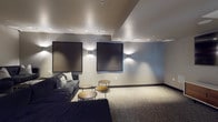 Located in the Arts District of Los Angeles at 950 E Third St, this single-floor screening room and lounge is designed for an immersive, cinema-inspired experience. The space is anchored by a large wall-mounted flat-screen display paired with a sleek, modern media console featuring warm wood accents and white cabinetry, providing a polished focal point for the room. Seating is arranged in a tiered, theater-style layout with multiple deep, plush dark sofas accented by decorative patterned pillows, ensuring clear sightlines from every position. Clustered round side tables in a brushed gold finish are thoughtfully placed throughout for added convenience. The walls are adorned with large decorative acoustic panels, enhancing both the aesthetic and the audio environment of the space. Ambient wall sconces cast a warm, dramatic uplight throughout, complementing the recessed ceiling lighting to create an intimate atmosphere. The floor is covered in neutral, textured carpet that adds comfort underfoot. A bold double-door entry with circular porthole windows serves as a distinctive architectural feature, offering a dramatic entrance into this well-appointed screening and entertainment lounge.