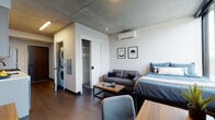 This thoughtfully designed studio showcases a seamless open-concept layout that combines living, sleeping, and cooking areas into one cohesive, efficient space. The architectural design is defined by striking exposed concrete ceilings and structural columns, lending an industrial-chic character balanced by clean white walls and warm dark hardwood flooring throughout.

The kitchen is outfitted with stainless steel appliances, including a gas range, over-the-range microwave, and dishwasher. Sleek, flat-front cabinetry in a soft neutral tone is complemented by open shelving and a crisp white countertop, with a matte black faucet adding a modern accent.

The main living area comfortably accommodates a bed, sofa, coffee table, and dedicated workspace with a desk and chair. A wall-mounted flat-screen TV and a ductless mini-split air conditioning unit add to the unit's practical conveniences. In-unit stacked washer and dryer are neatly tucked into a closet alongside additional storage.

Floor-to-ceiling windows flood the sleeping area with natural light and frame expansive panoramic city views, creating a bright and airy atmosphere. The bathroom features a walk-in glass-enclosed shower with large-format grey tile surround and matte black hardware, delivering a clean, contemporary finish throughout.