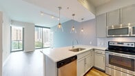 This well-appointed 2-bedroom, 2-bathroom residence offers a thoughtfully designed single-level layout with contemporary finishes and an abundance of natural light. The open-concept living area is anchored by expansive floor-to-ceiling windows that frame sweeping urban skyline views, allowing natural light to flood the space across wood-style flooring.

The kitchen serves as a functional centerpiece, featuring a large peninsula with quartz countertops, an undermount sink, and striking glass pendant lighting. Gray shaker-style cabinetry, subway tile backsplash, and a full suite of stainless steel appliances — including a French door refrigerator, electric range, built-in microwave, and dishwasher — complete the space with a polished, modern aesthetic.

The primary bedroom is a comfortable retreat with direct access to a private balcony through floor-to-ceiling sliding glass doors, offering additional city views. The en-suite bathroom includes a soaking tub with tile surround, a vanity with quartz countertops, and access to a dedicated walk-in closet with wire shelving. The second bedroom is bright and versatile, with its own access to a closet. Both bathrooms are finished with tile flooring and gray cabinetry. Wood-style flooring and a neutral color palette unify the space throughout.