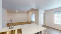 This single-floor residence is designed with a clean, modern aesthetic highlighted by a neutral color palette and an open, functional layout. The kitchen serves as a central hub, featuring white shaker-style cabinetry with brushed nickel hardware, light quartz countertops, stainless steel appliances including a refrigerator, microwave, and range, and a peninsula with an integrated dishwasher that opens to the living area. The kitchen's wood-look flooring extends into the adjacent dining and living spaces, which also include a dedicated exterior door providing convenient outdoor access and multiple large windows that bring in generous natural light.

The living area transitions to carpeted flooring and features vaulted ceilings with recessed lighting, lending an airy, open feel to the space. A dedicated laundry area with a full-size washer and dryer is conveniently located off the main living space. Both bedrooms are carpeted and well-lit by natural light, with one featuring a walk-in closet with built-in shelving. The two bathrooms are finished with warm stone-toned tile flooring and white vanities; one includes a shower-tub combination, while the other offers a standalone vanity and toilet configuration.